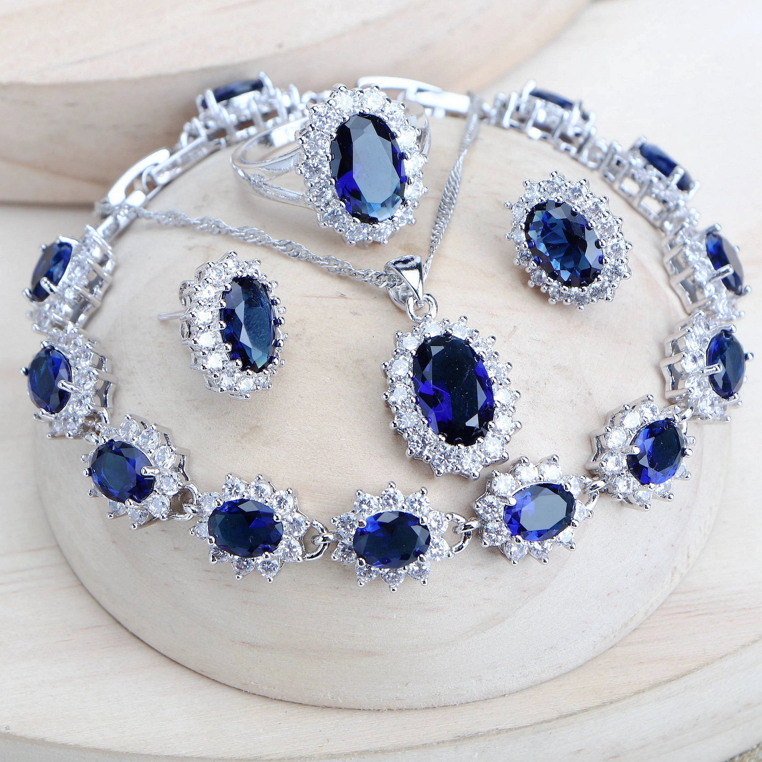 A matching jewelry set featuring a silver-tone bracelet, necklace, earrings, and ring, all adorned with deep blue oval gemstones and surrounded by clear rhinestone-like accents, displayed on a light wood surface.