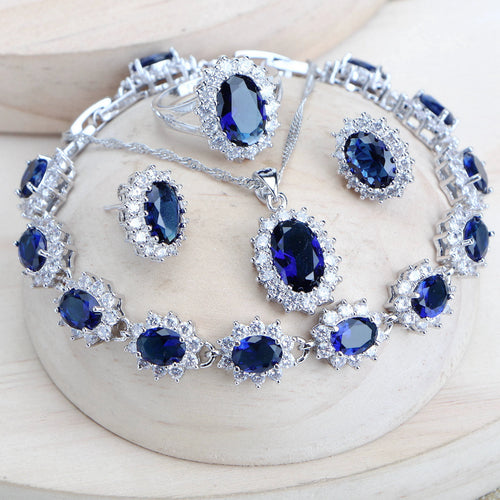 A matching jewelry set featuring a silver-tone bracelet, necklace, earrings, and ring, all adorned with deep blue oval gemstones and surrounded by clear rhinestone-like accents, displayed on a light wood surface.
